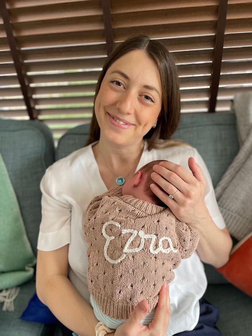 Woman holding a baby wrapped in a pink blanket with 'Ezra' on it, sitting on a couch.