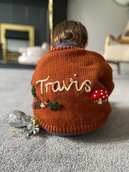 Child wearing a brown sweater with 'Travis' embroidered on it, sitting on a carpeted floor.