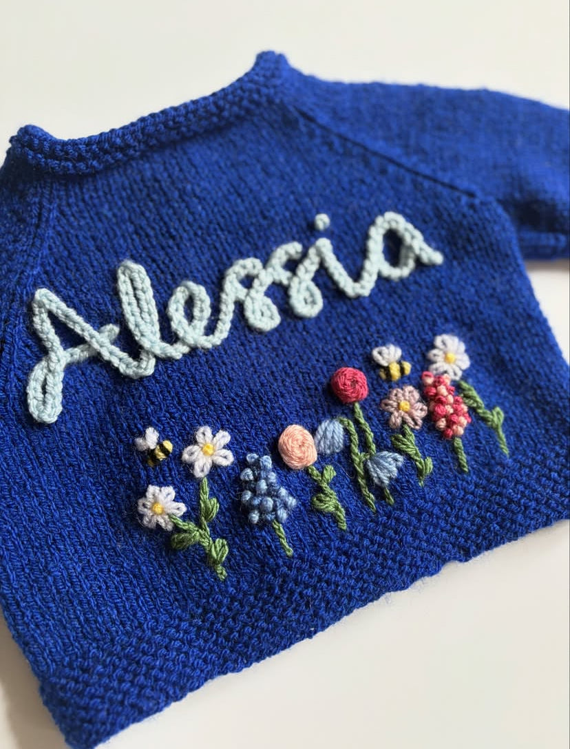 Blue knitted sweater with embroidered name 'Alessia' and colorful flowers on a white background