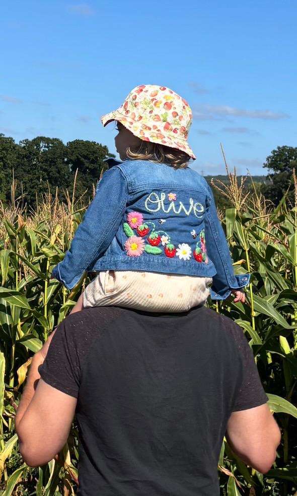 Person holding a child in a cornfield, with the child wearing a denim jacket with floral embroidery and 'Olivia' written on it.
