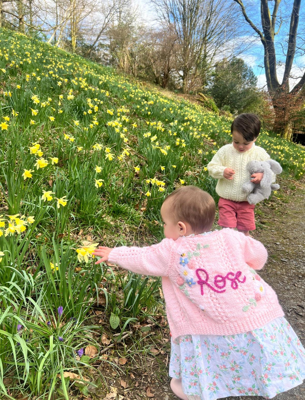 Two children in a field of daffodils, one wearing a pink coat with 'Rose' on it.
