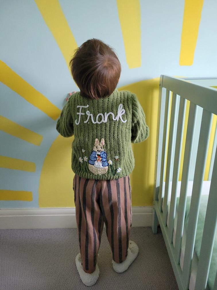 Child wearing a green sweater with 'Frank' on it, standing in a room with a crib and sun design on the wall.